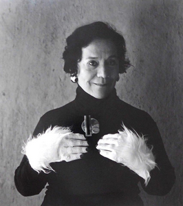 Autorretrato de la fotógrafa Flor Garduño, vestida de negro con adornos blancos en las muñecas, sus manos se encuentran sobre su pecho. La fotógrafa sonríe al espectador con complicidad.